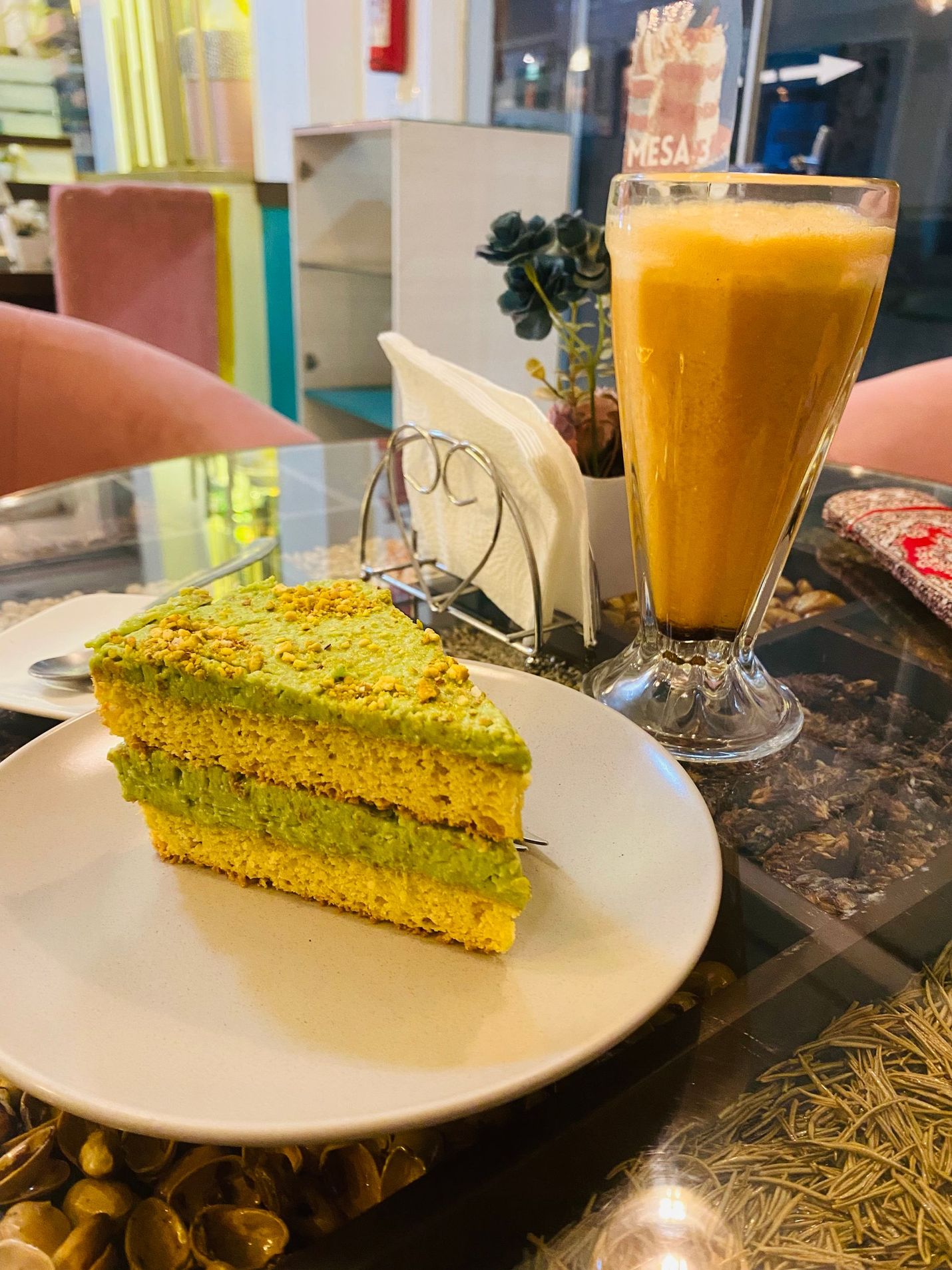 Slice of green cake on a plate beside a tall orange smoothie in a café