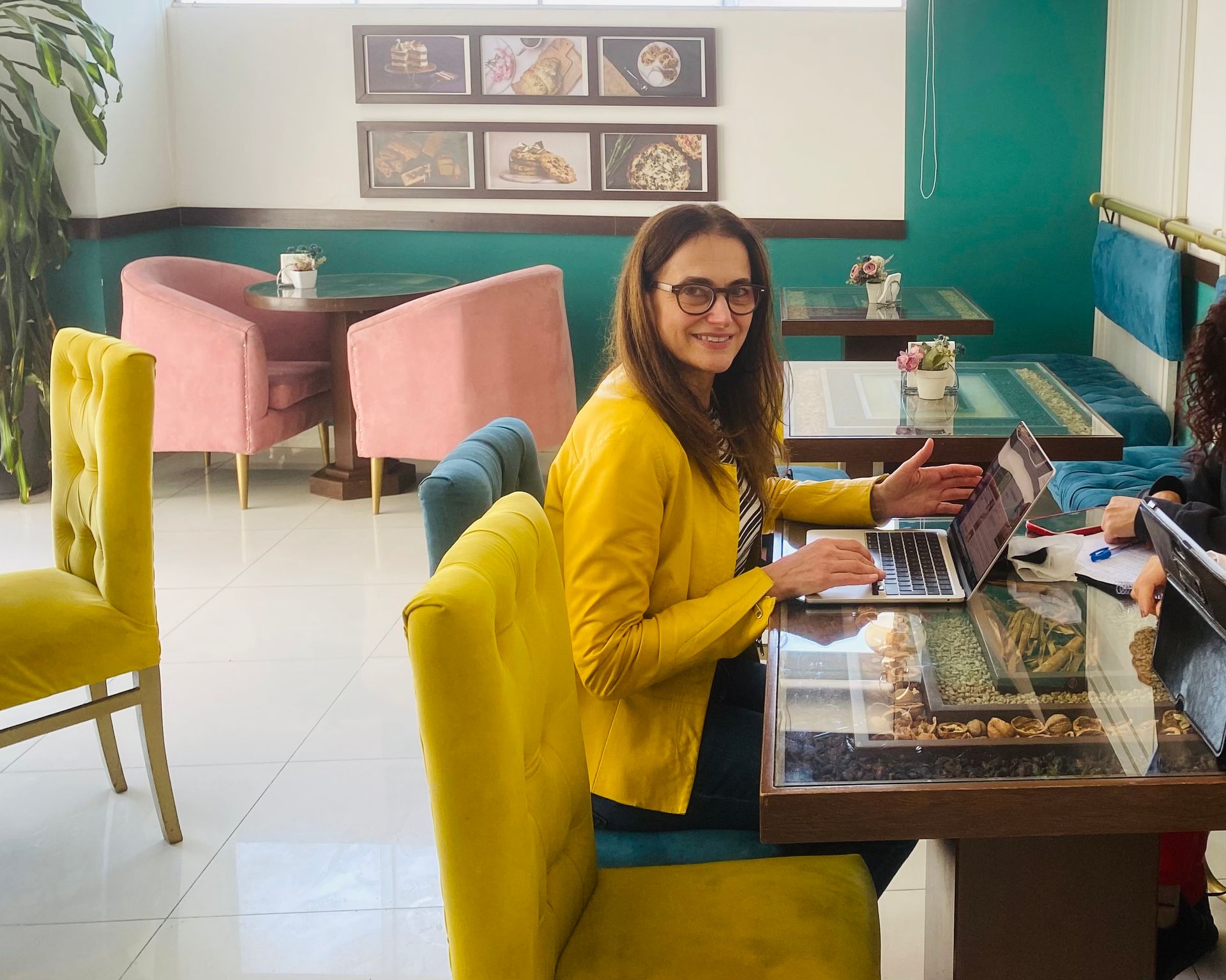 Woman in yellow jacket smiling at a laptop in a colorful cafe booth