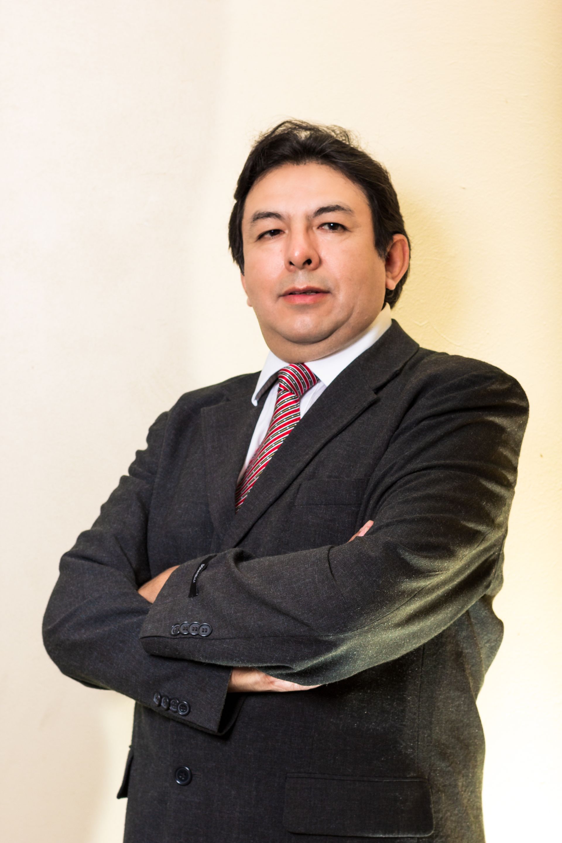 Un hombre con traje oscuro y corbata a rayas rojas está de pie con los brazos cruzados contra un fondo claro.