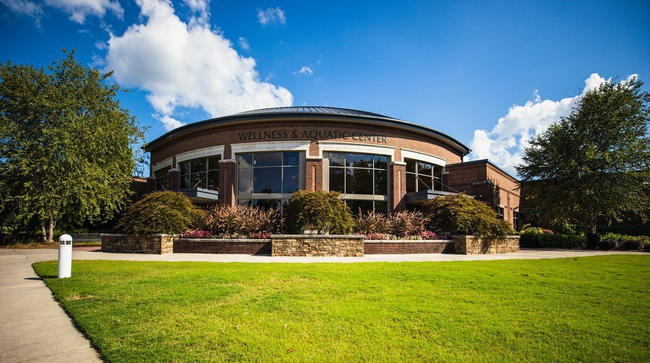 Brick building of Cullman Wellness and Aquatic Center with a curved roof and large windows, fronted by a green lawn and trees under a blue sky.