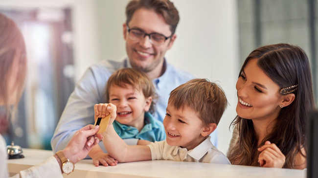 Family smiling at a table, with two children holding a room key while adults look on in a bright room