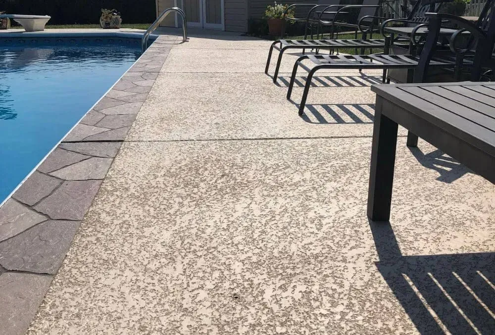 Concrete pool deck beside blue swimming pool with lounge chairs and a patio table in sunlight