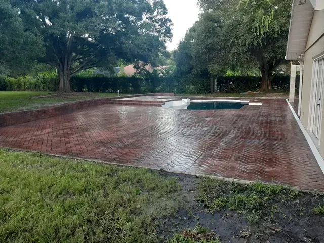 Rain-soaked brick patio beside a house, with a small pool and grassy yard bordered by trees.