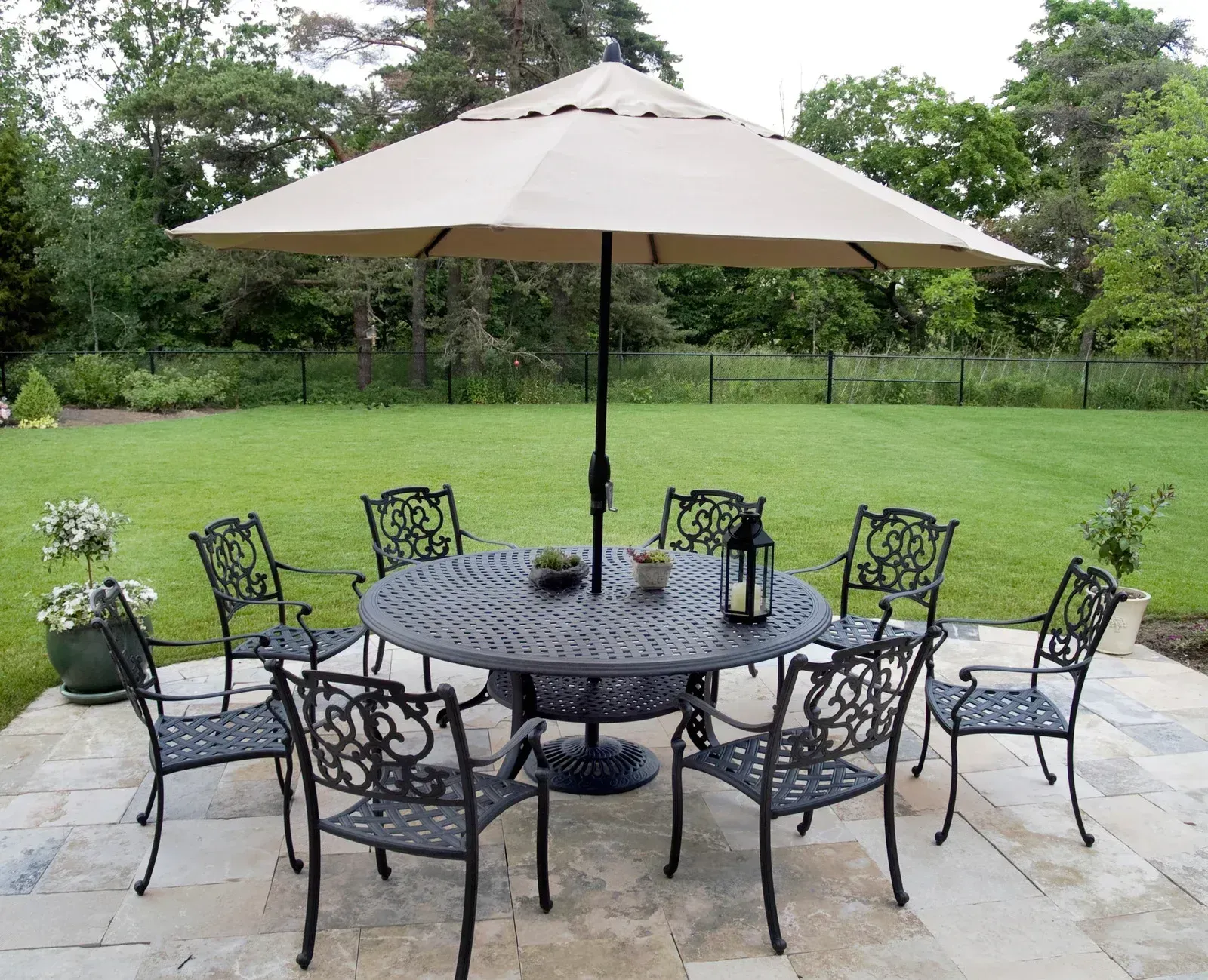 Patio dining set with black chairs and beige umbrella on stone pavers overlooking a green lawn