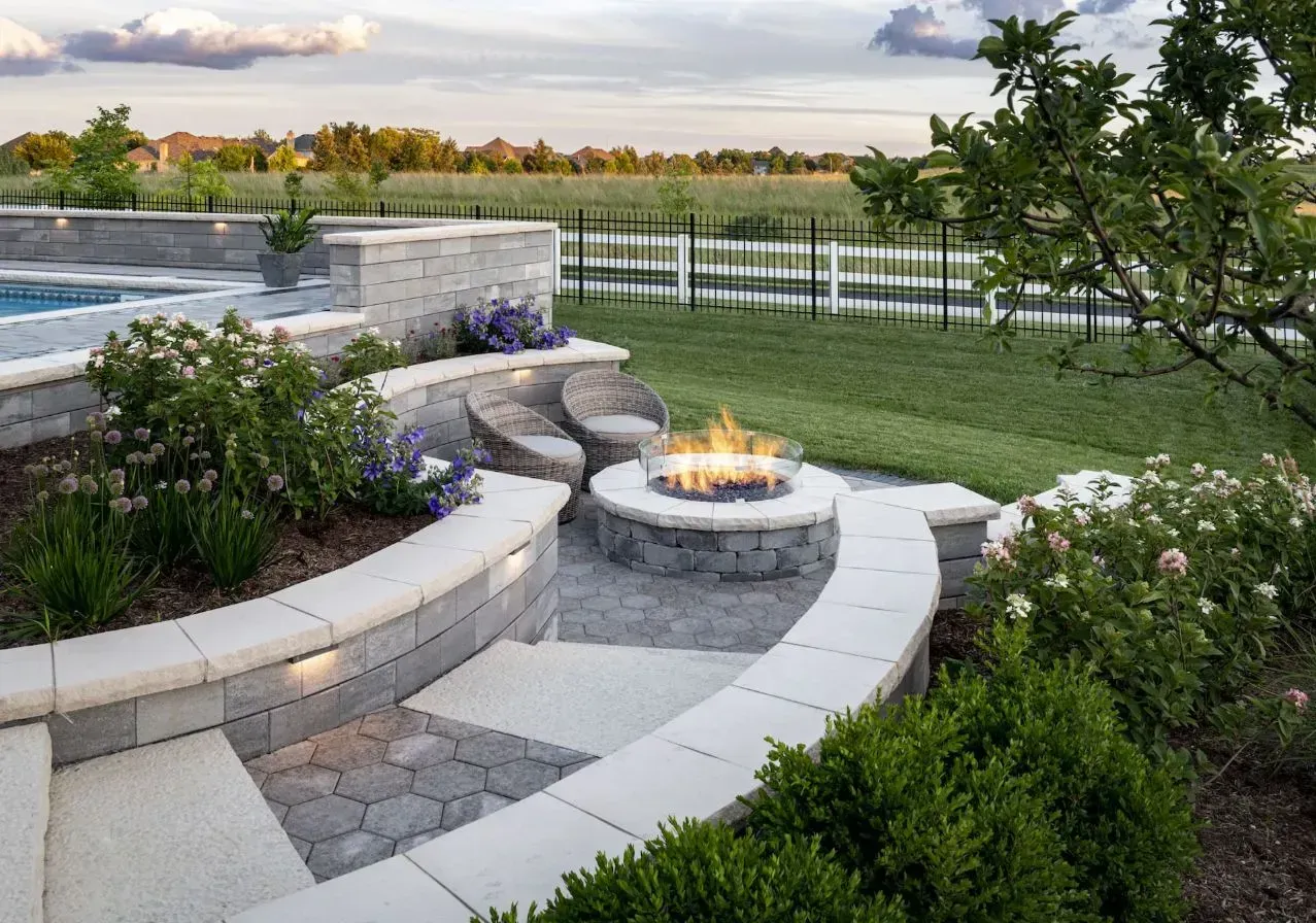 Backyard patio with curved stone fire pit, pool edge, and landscaped garden at sunset