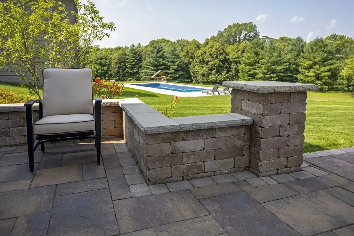 Patio chairs beside a stone fire pit overlooking a backyard pool and lawn