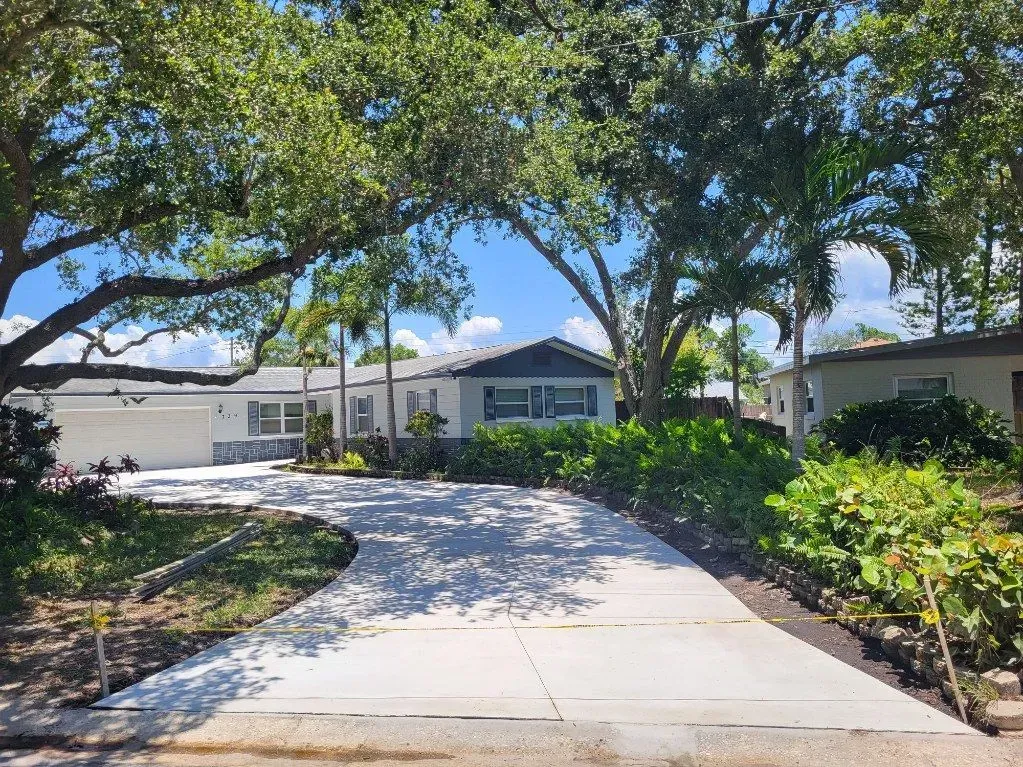 Tree-lined driveway leading to a white house with a landscaped front yard and sunny blue sky