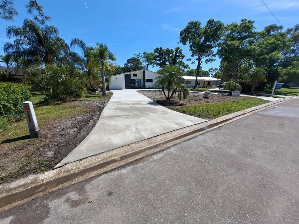 Suburban driveway leading to a small white house with palm trees and a yard under a clear blue sky