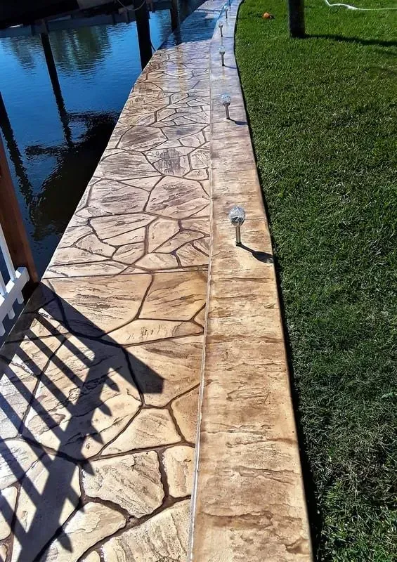 Stamped concrete walkway beside a pool and green lawn, with shadows from a fence.