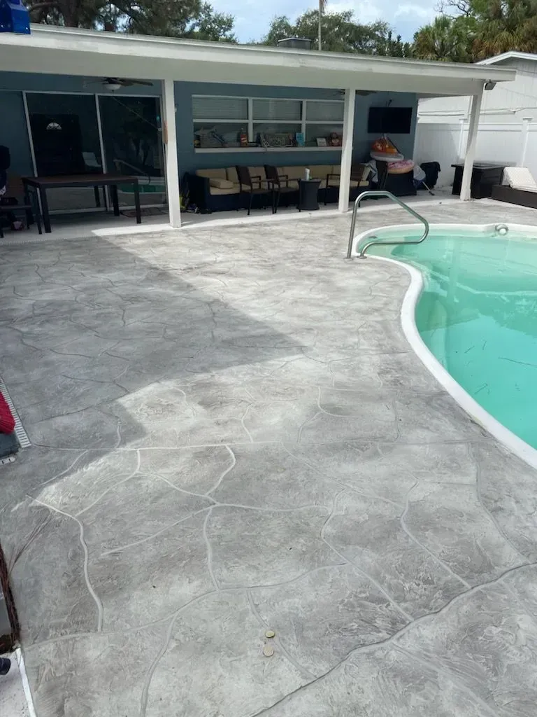 Concrete pool deck beside a turquoise swimming pool and covered patio with seating and a grill