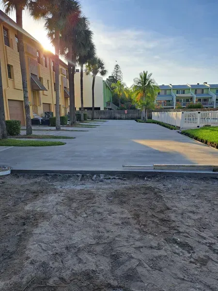 Sunlit walkway beside beige apartment buildings, palm trees, and a white fence near the beach