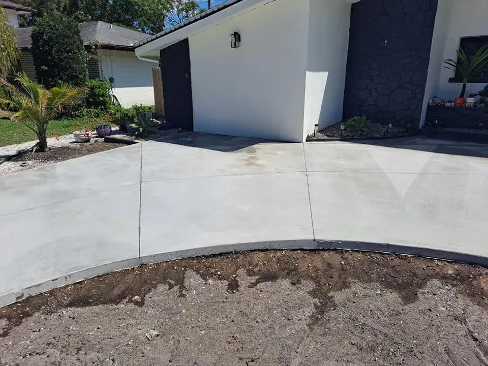 Curved concrete driveway beside a white house and dark garage, with a bare dirt strip in front.