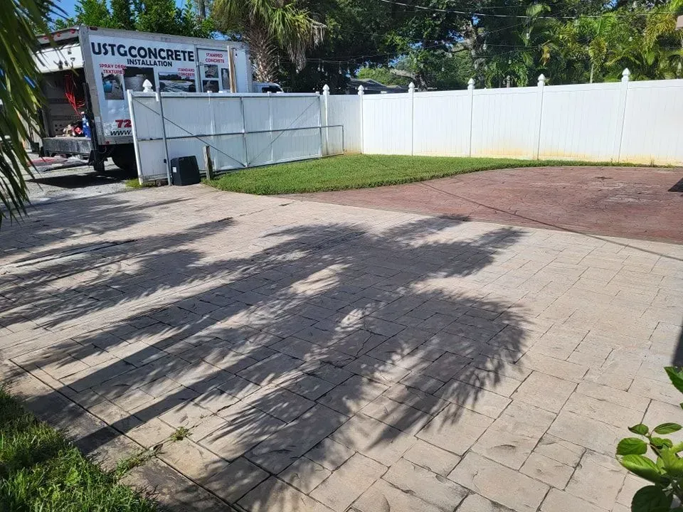 Driveway beside a white fence and brick patio, with tree shadows and a truck in the background