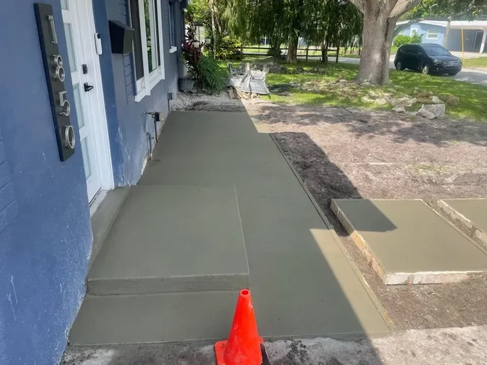 Fresh concrete front walkway beside a blue house, with an orange traffic cone and yard in the background