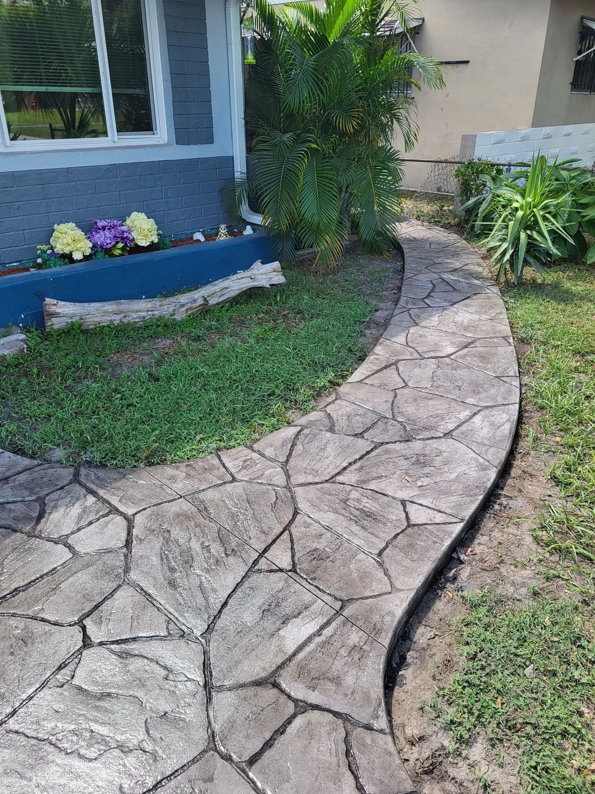 Curved stone walkway beside a house, with lawn, shrubs, and flower bed.