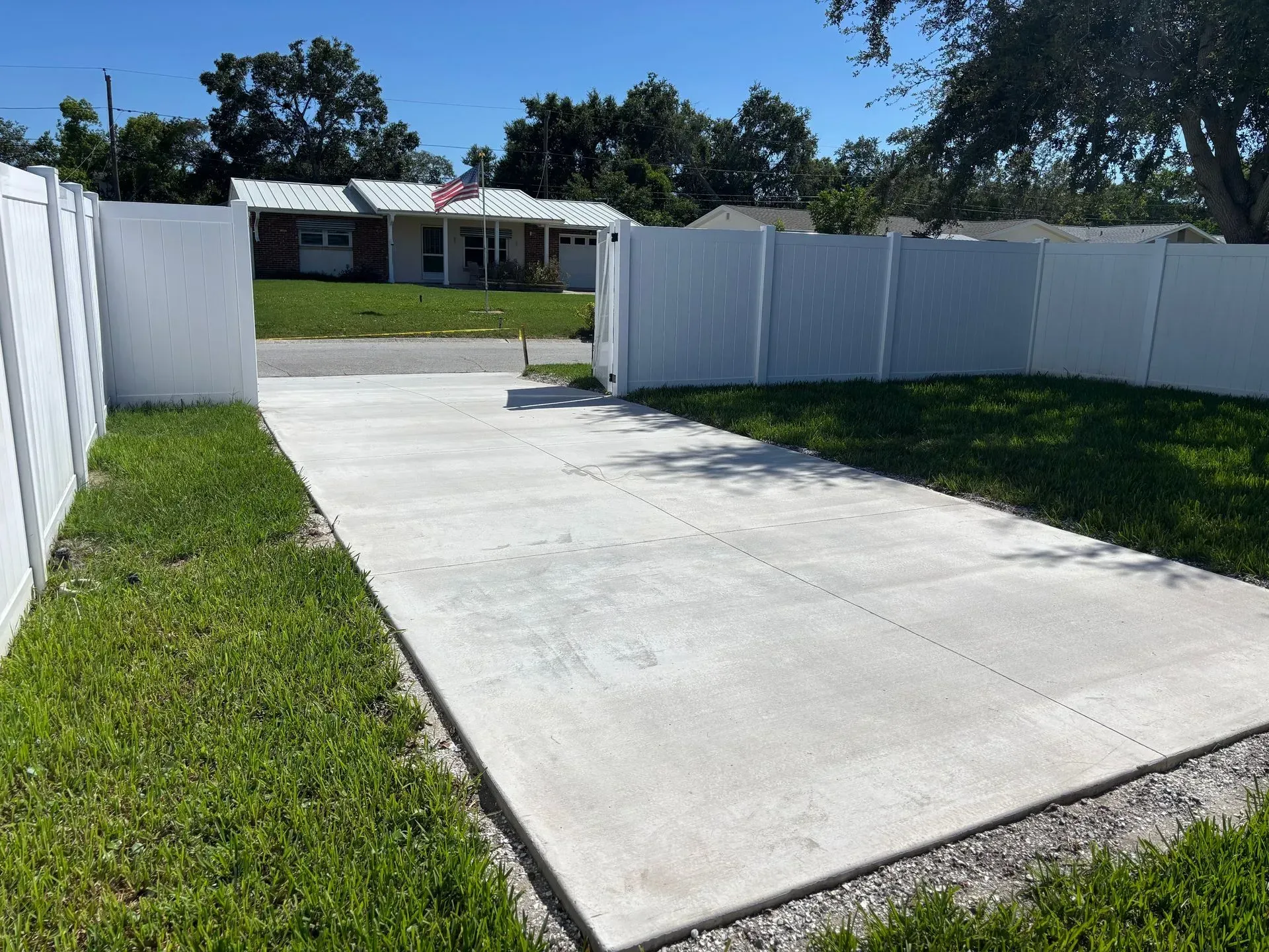 Concrete driveway between white fence gates leading to a single-story house with a lawn and trees.