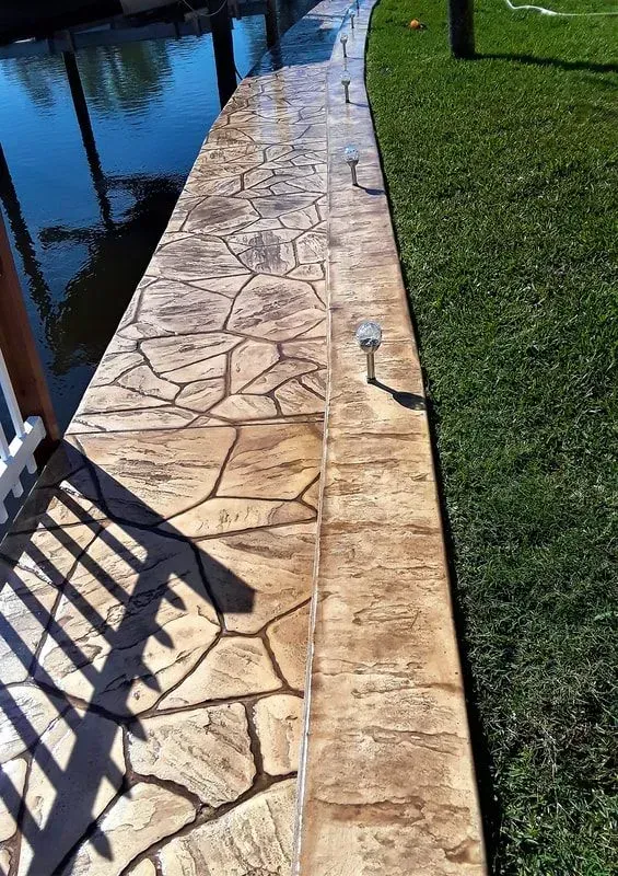 Stone poolside walkway beside blue water and green grass, with a white railing shadow.