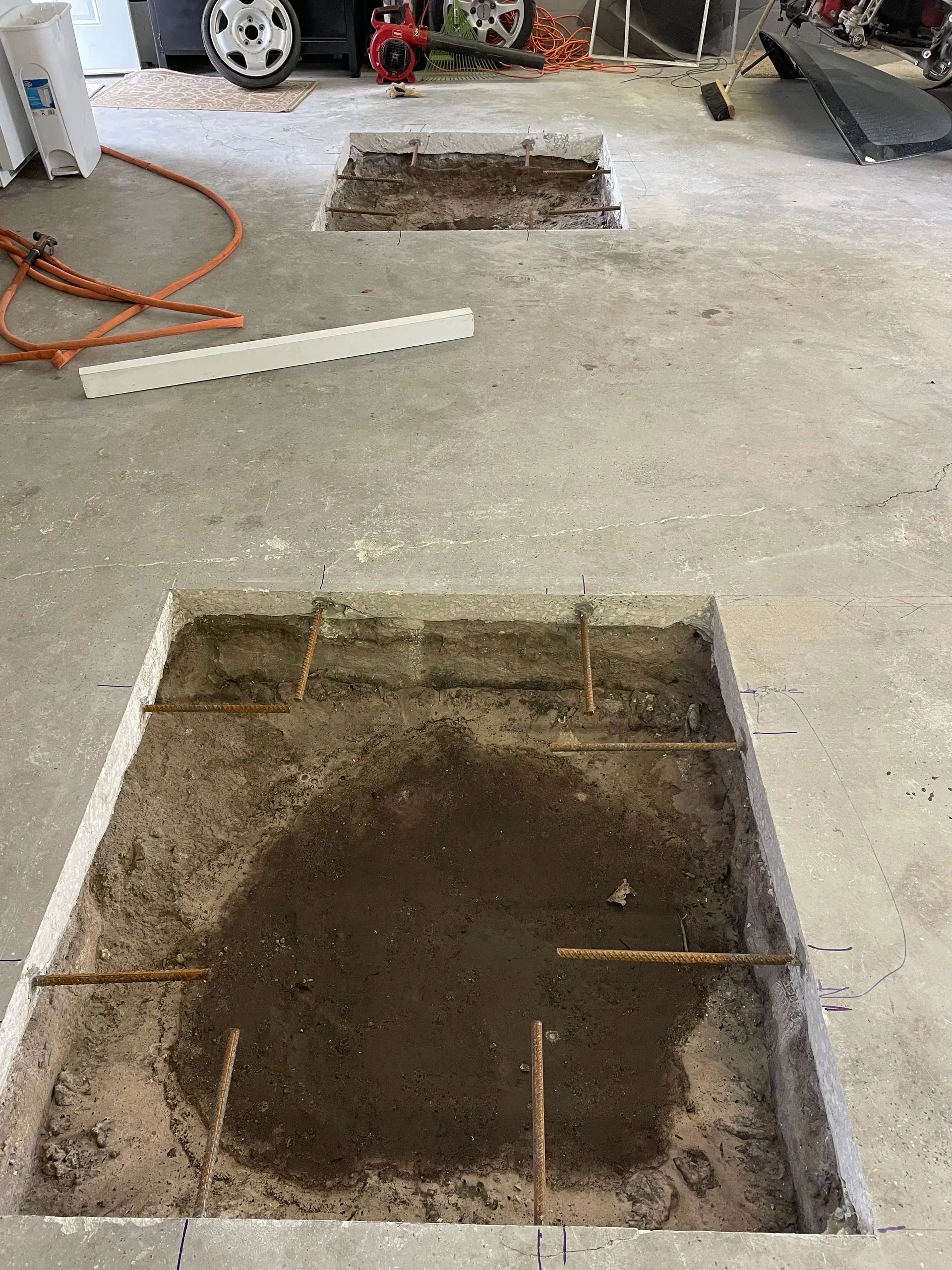 Concrete floor with two square excavation pits in a garage-like space, one covered by a white plank and orange hose nearby
