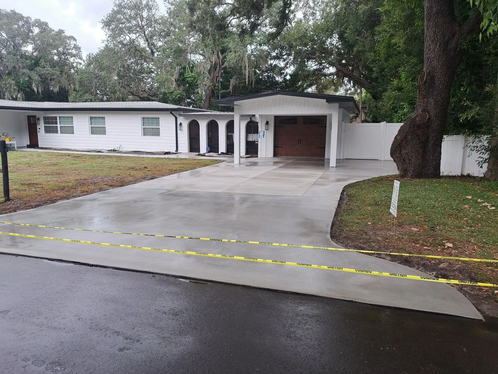 Driveway leading to a single-story white house with a garage, wet pavement, and yellow caution tape across the entrance