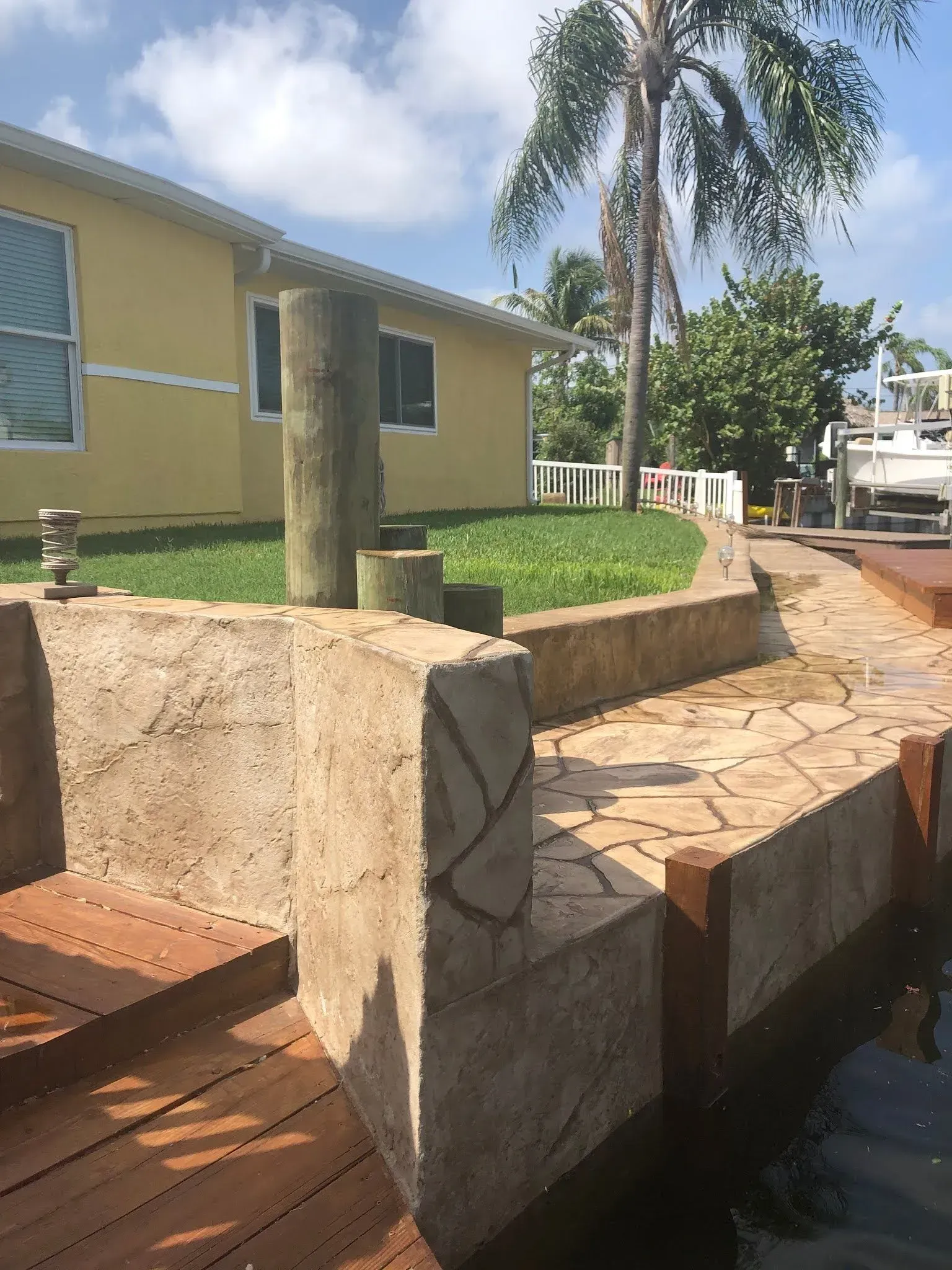 Stone walkway beside a yellow building and canal, with palm trees and a wooden bench in the foreground.