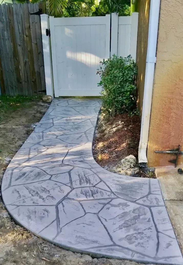 Curved stamped-concrete walkway leading to a white gate beside a tan wall and small shrub