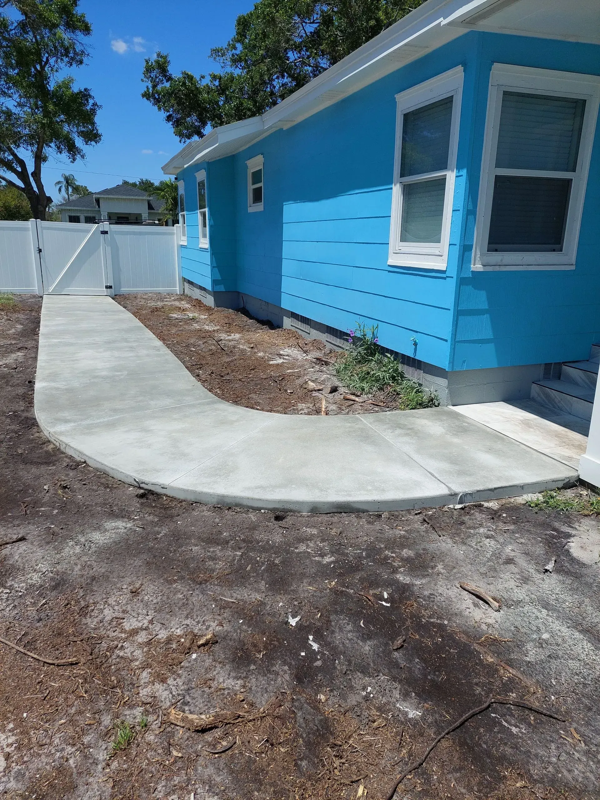 Blue house exterior with curved concrete walkway, white fence, and mulch bed under a clear sky