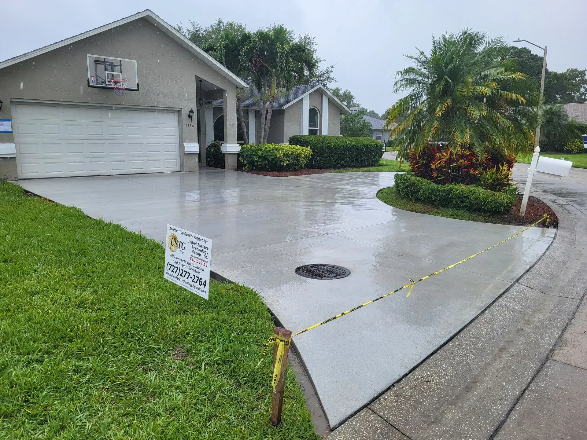 Wet suburban driveway with a house, garage, palm tree, and a small yard sign near the curb