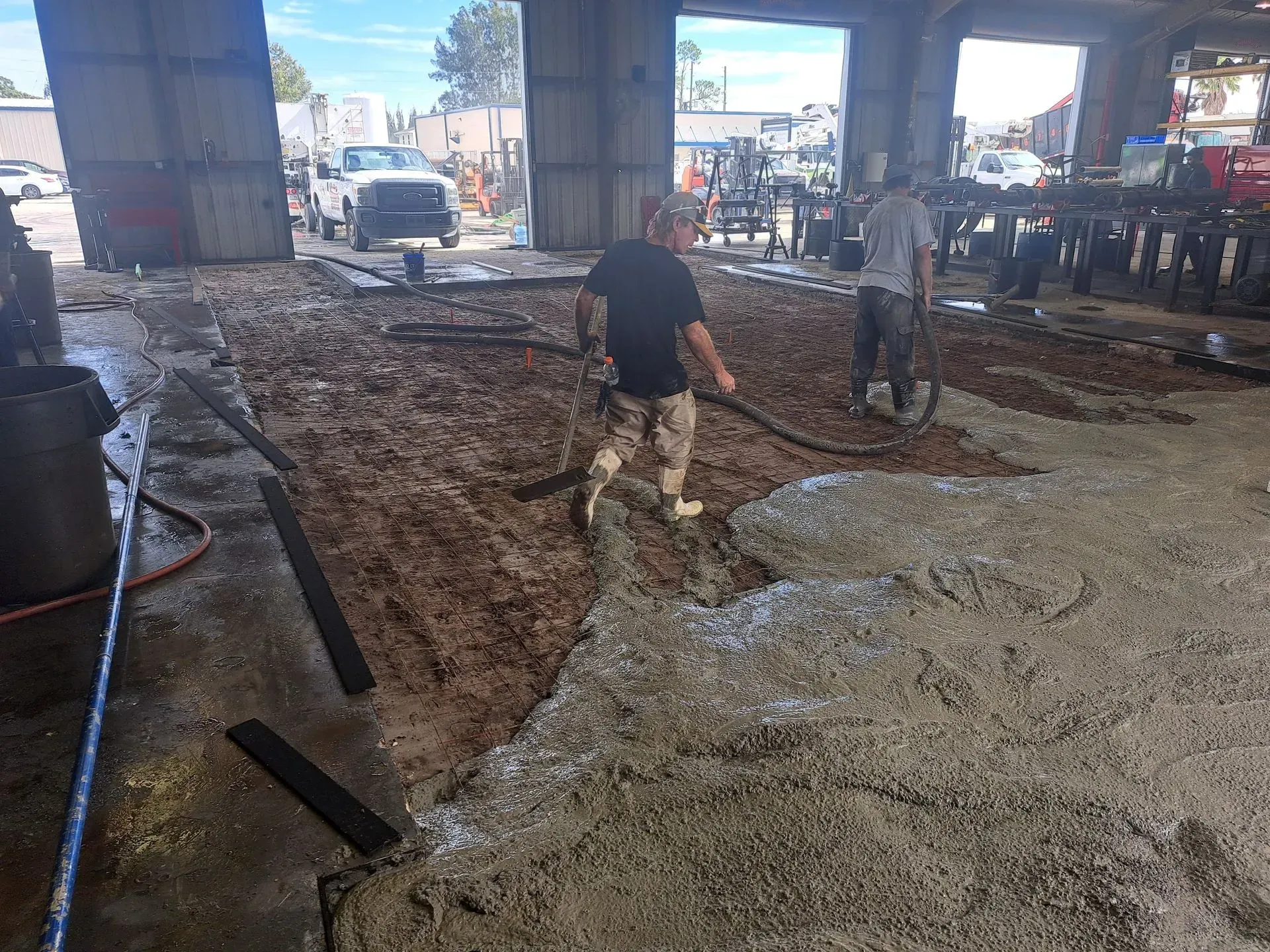 Construction workers walking through a muddy excavation under an overpass, with gravel piles and equipment nearby