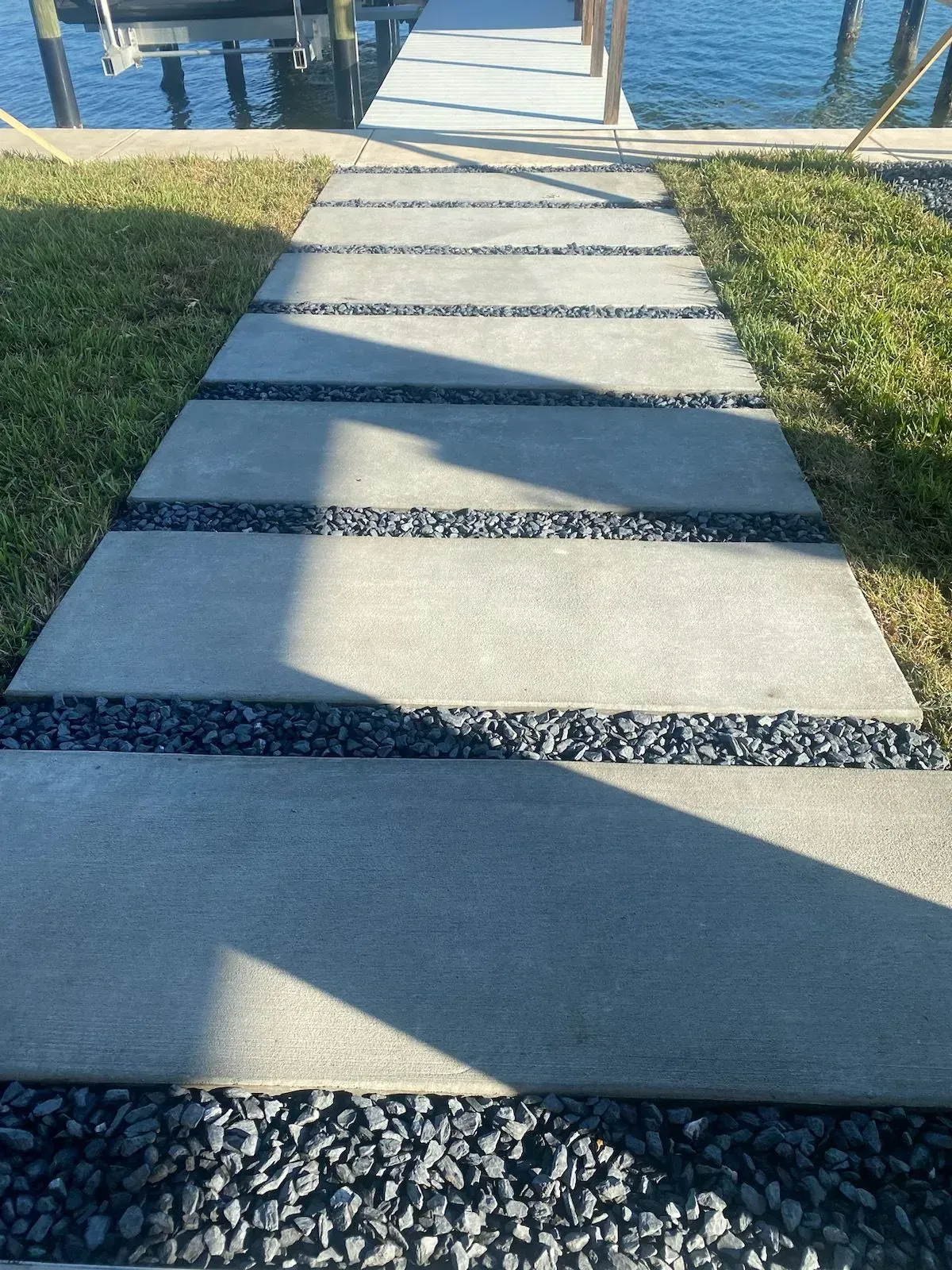 Concrete path with alternating stone slabs leading to water, bordered by grass and gravel, with sharp shadows.
