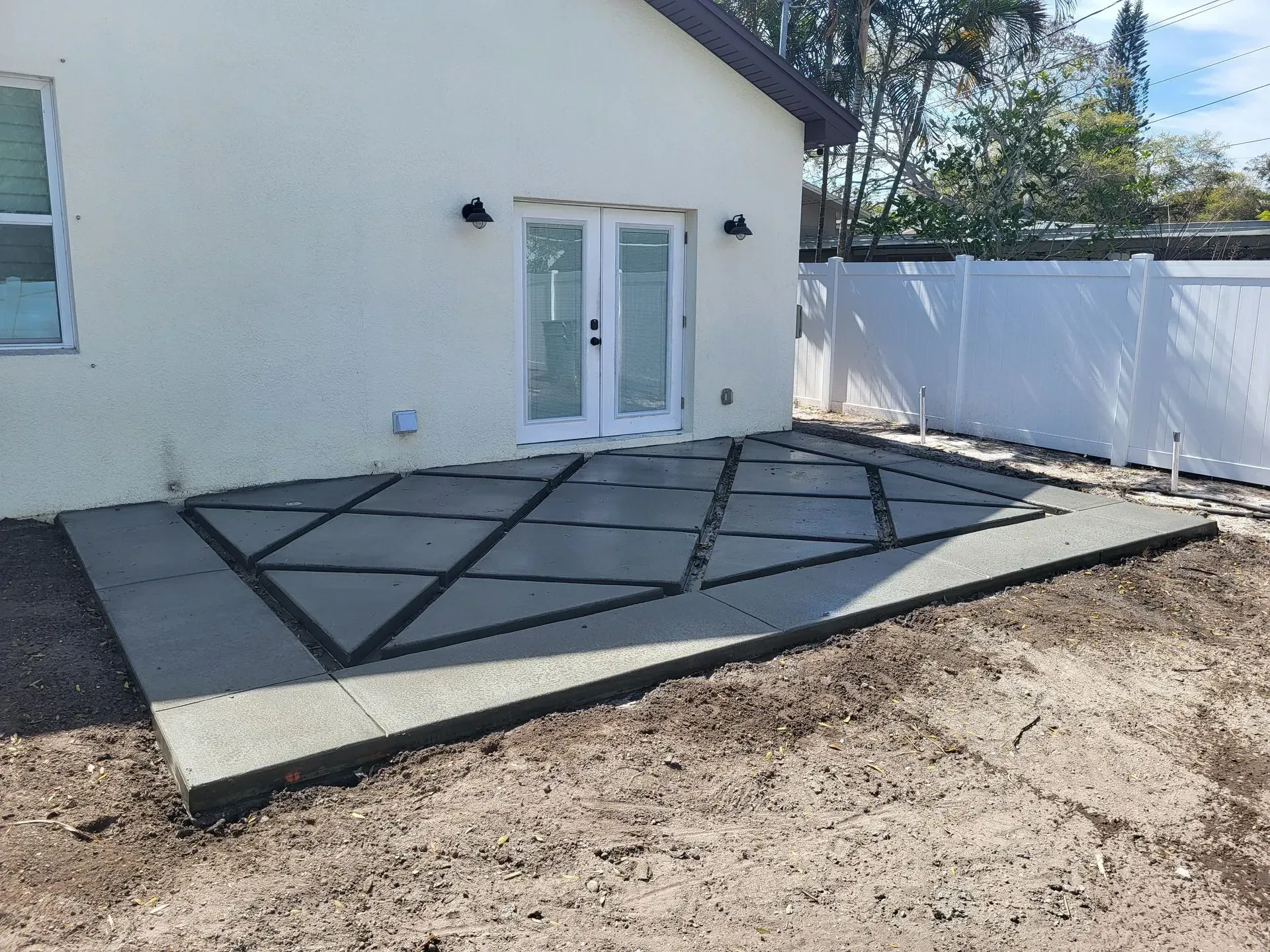 Concrete patio with black grid lines beside a house and white fence, under construction