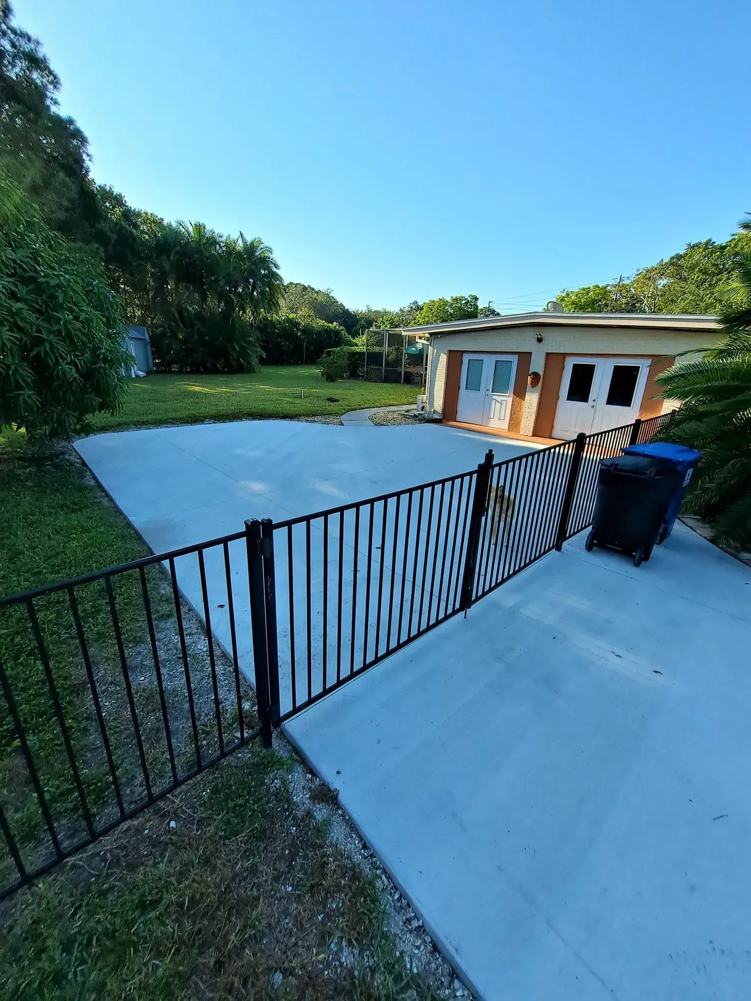Backyard patio with black fence, blue pavement, lawn, trees, and a small outbuilding under a clear sky