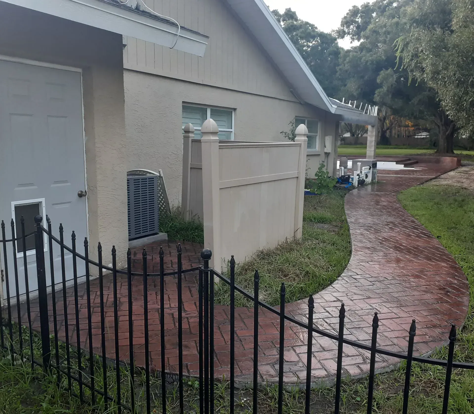 Rainy residential walkway beside a beige house, black fence, and brick path curving past trees.