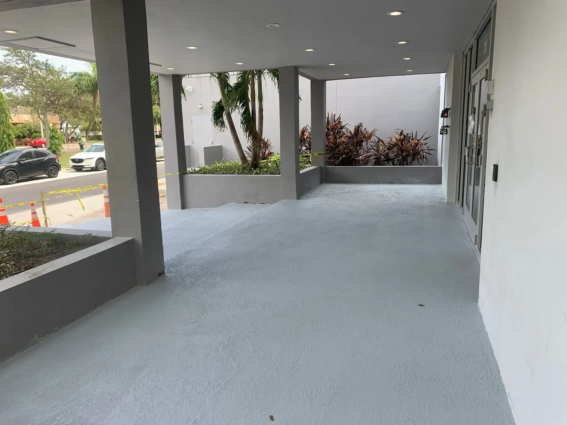 Covered walkway with gray floor, white columns, and planters along a building entrance