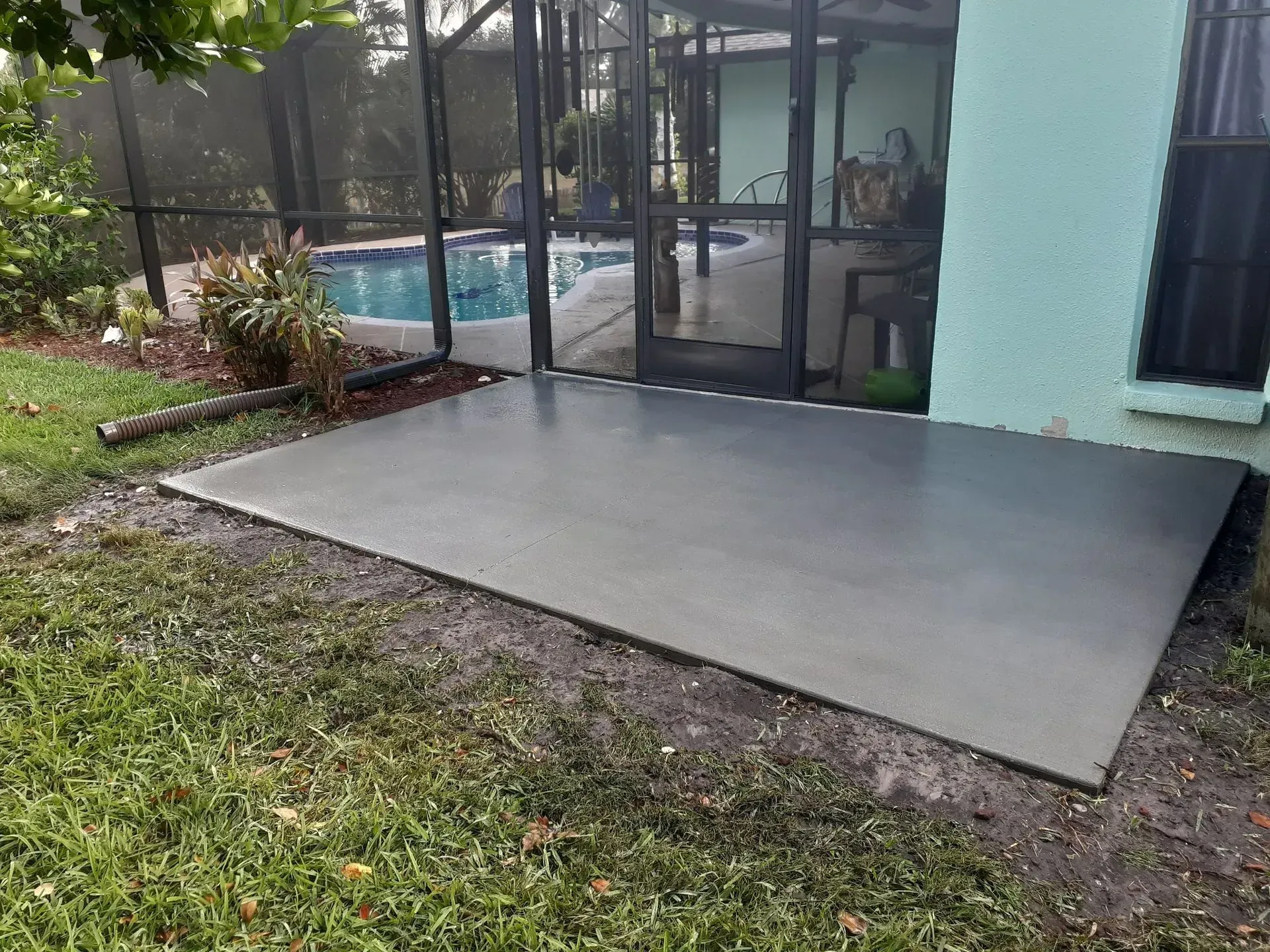 Concrete patio slab beside a screened pool enclosure, with grass and a turquoise house wall.