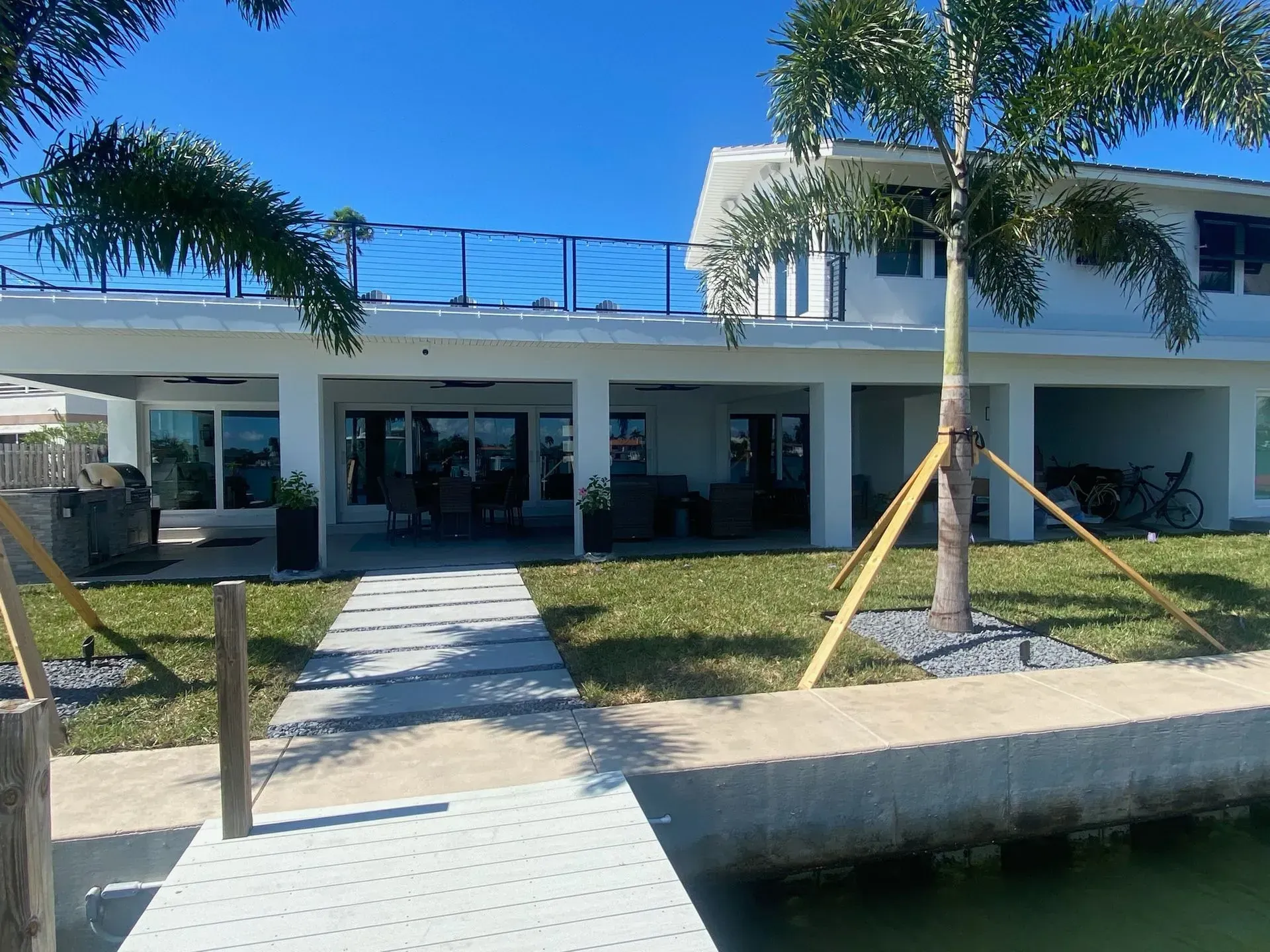 Modern white waterfront house with palm trees and a walkway over water under a clear blue sky