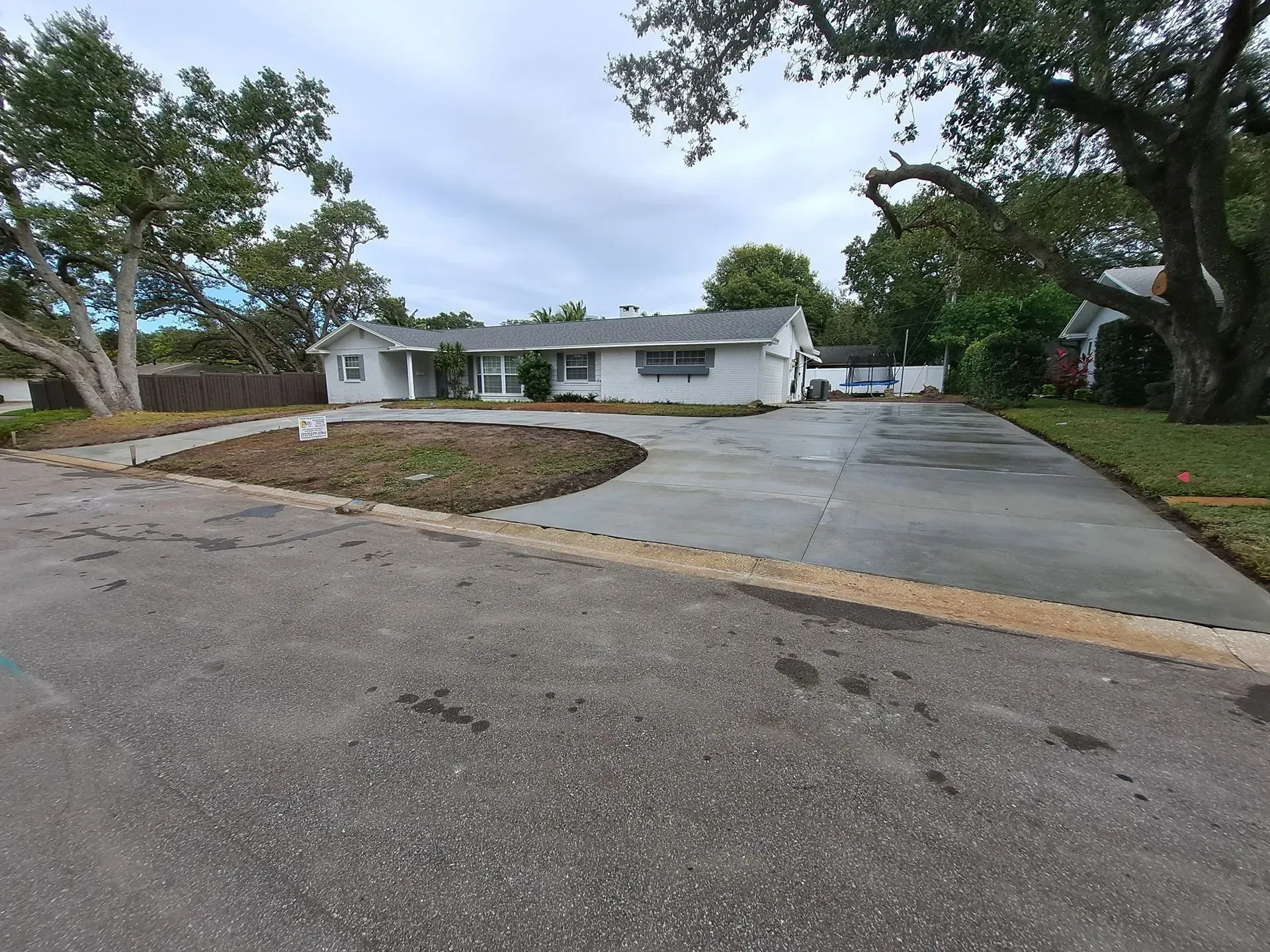 Quiet suburban street with a white house, wide driveway, and trees under a cloudy sky