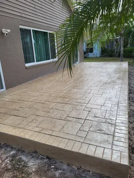 A tan stamped-concrete patio beside a house, with a raised border and palm fronds overhead.