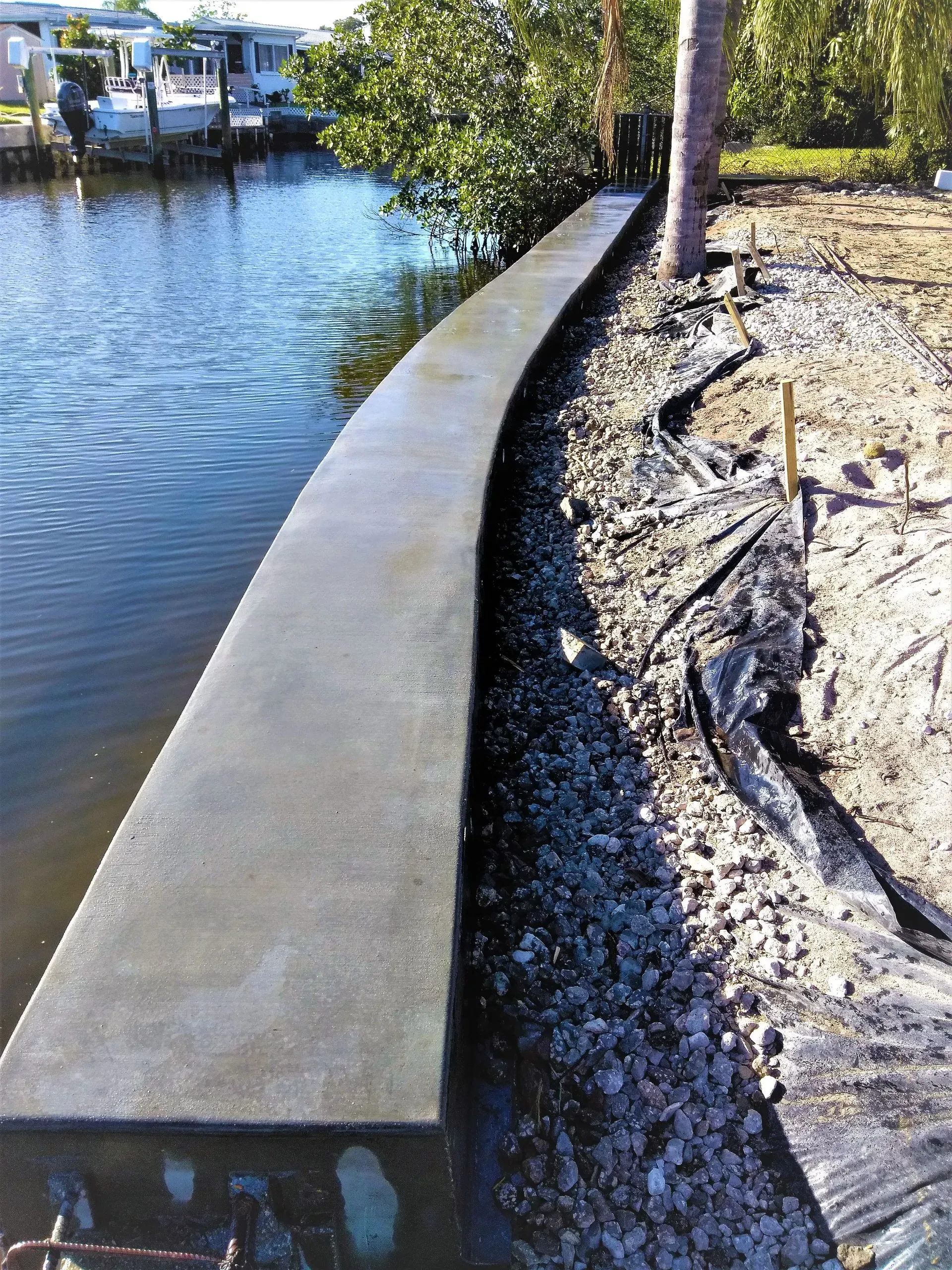 Concrete seawall along a calm waterfront beside rocks, trees, and a canal.