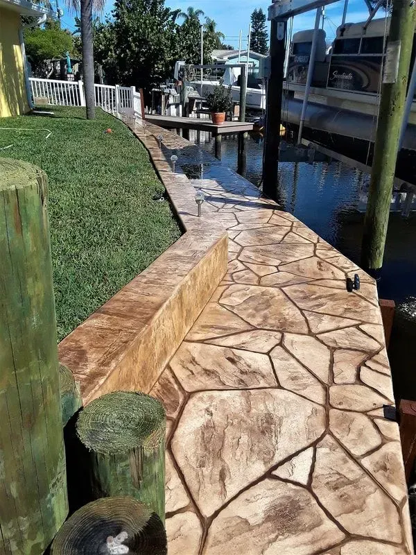 Stamped-concrete walkway beside a canal, with grass slope, dock, and boats moored along the right side.