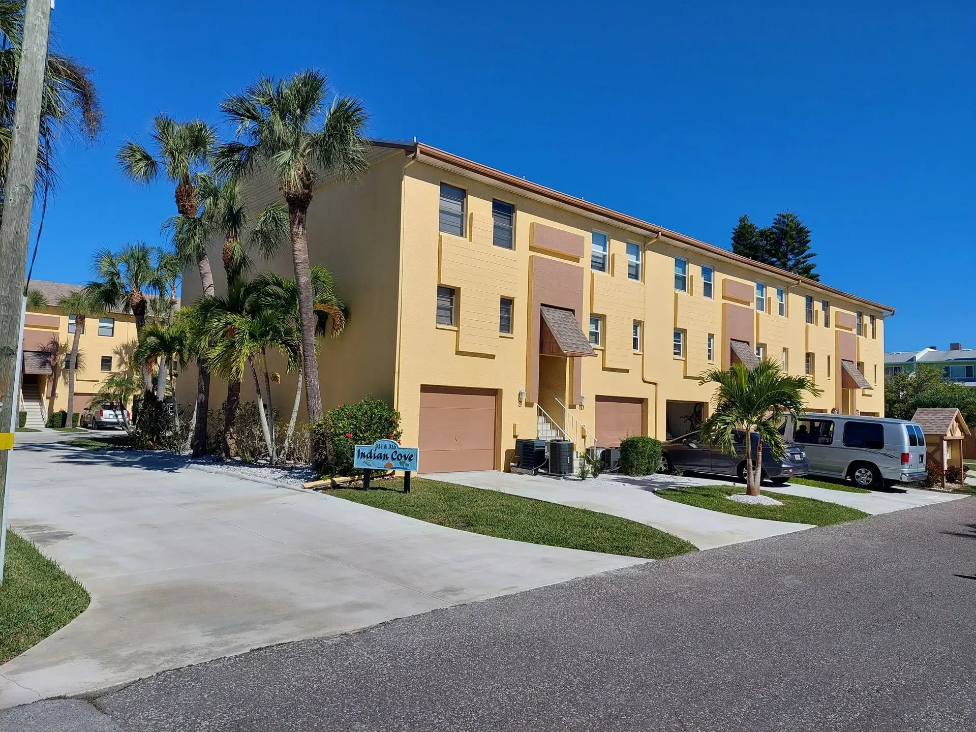 Yellow apartment building with palm trees, parked cars, and a driveway under a clear blue sky