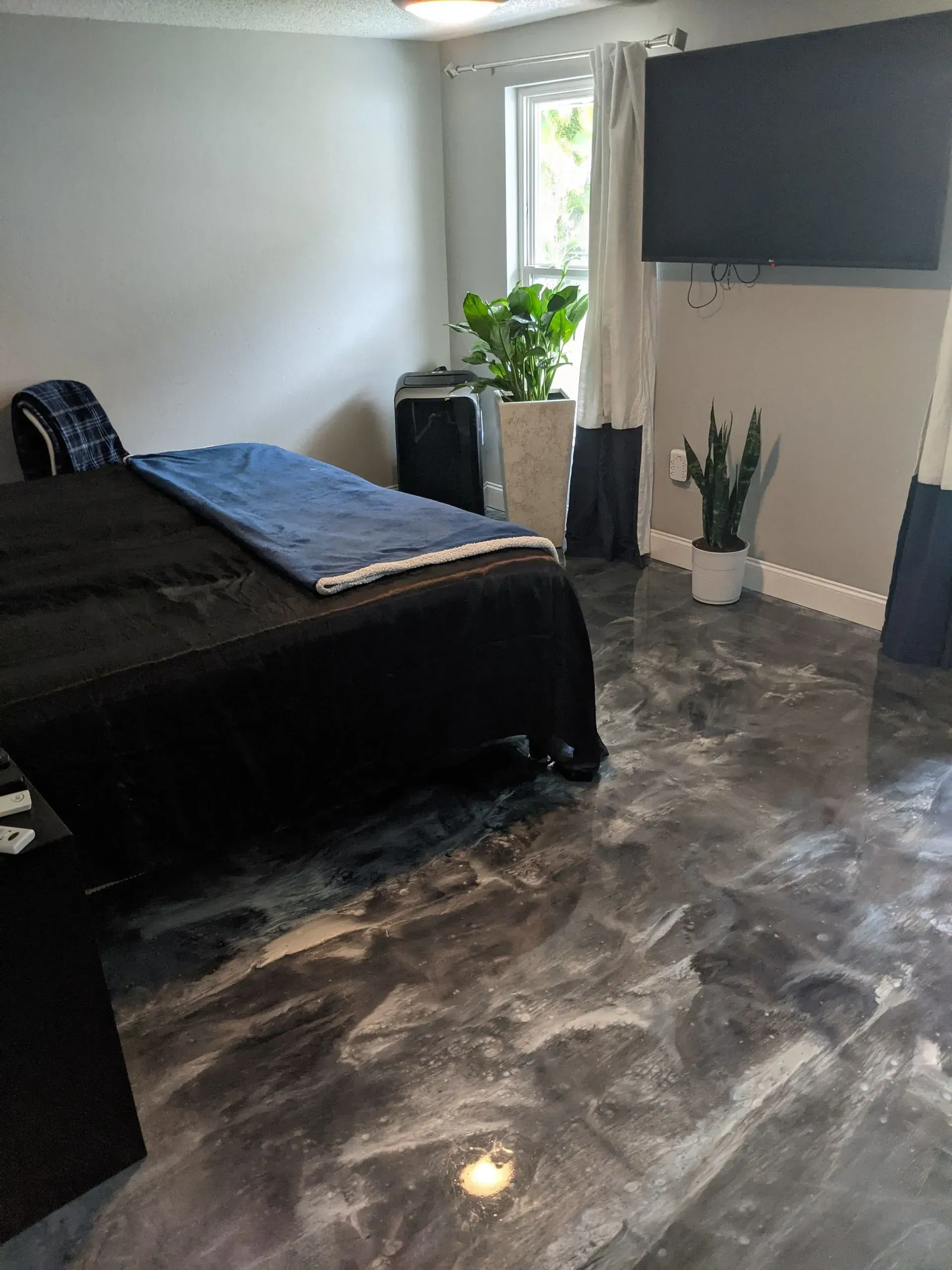Modern bedroom with black bed, glossy marble floor, wall-mounted TV, and plants by a window.