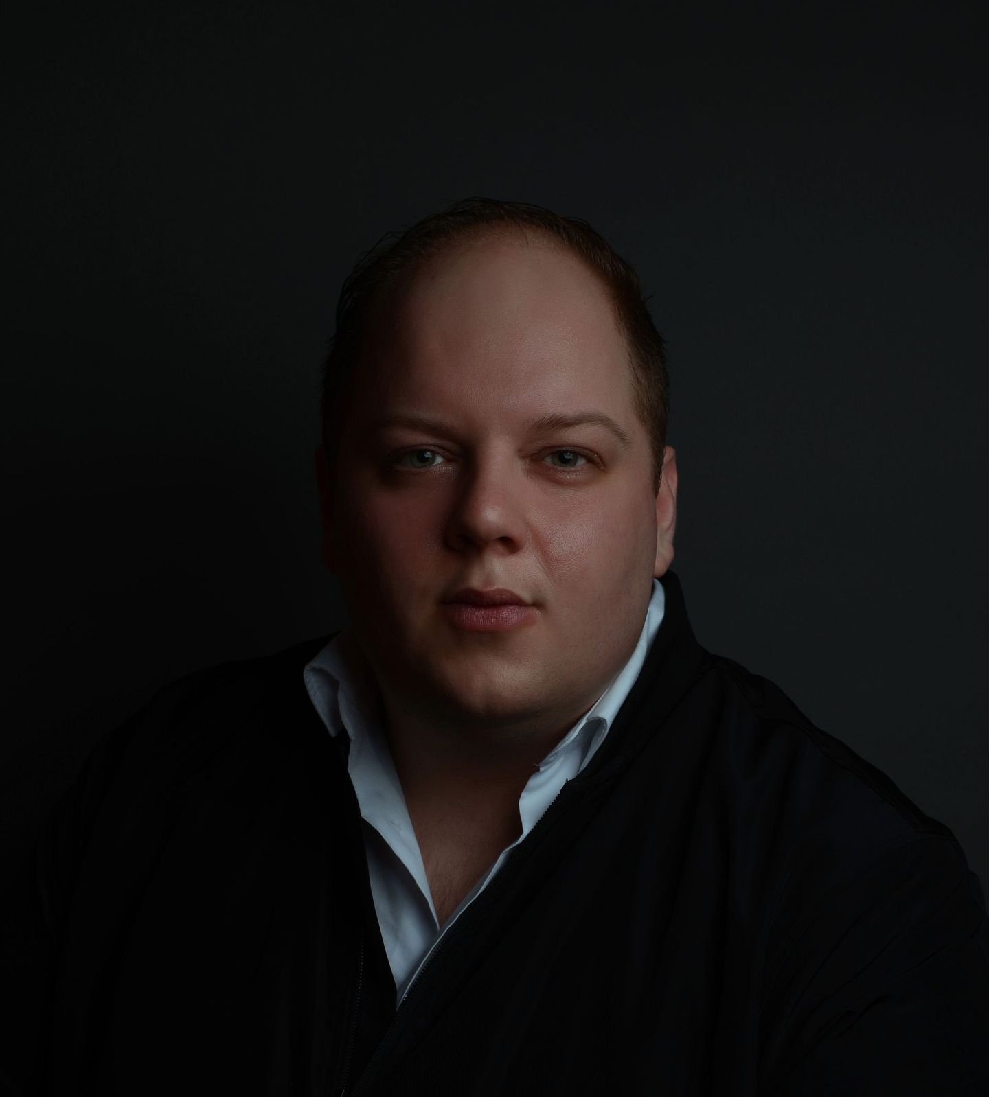 Een man in een zwarte trui en een wit overhemd kijkt naar de camera.