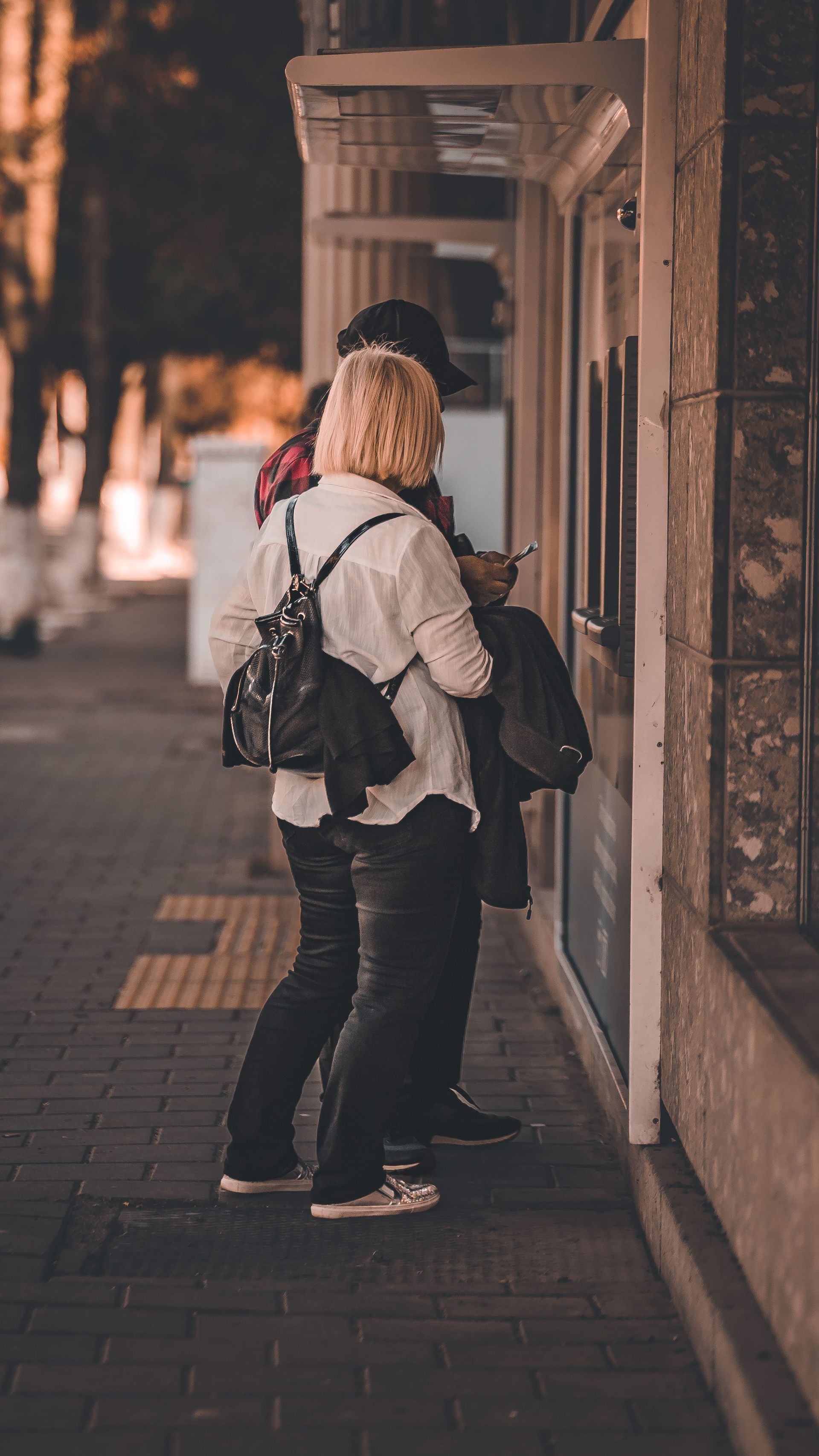Two people standing on a sidewalk looking into a shop window at dusk