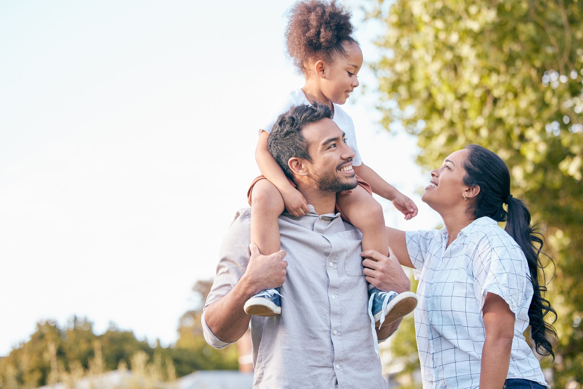 A man is carrying a little girl on his shoulders while a woman looks on.