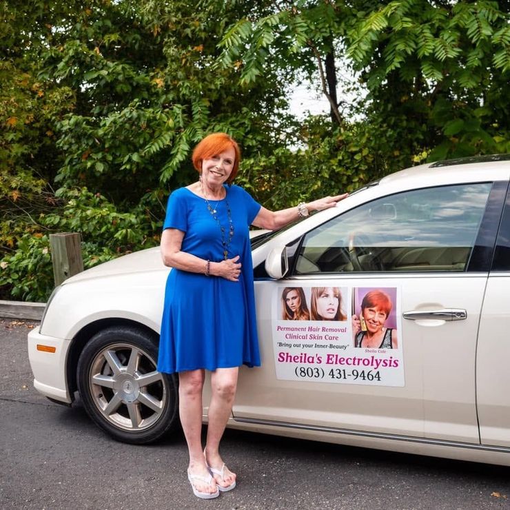 Woman in blue dress standing beside a white car with a “Sheila’s Electrolysis” sign on the door