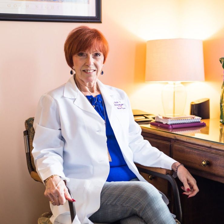 Doctor in a white coat seated at a desk beside a lit lamp, holding a cane and smiling.