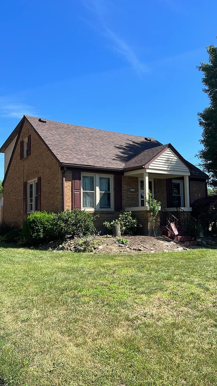 Brick house with brown roof and shutters, small front porch, surrounded by greenery, bright blue sky.
