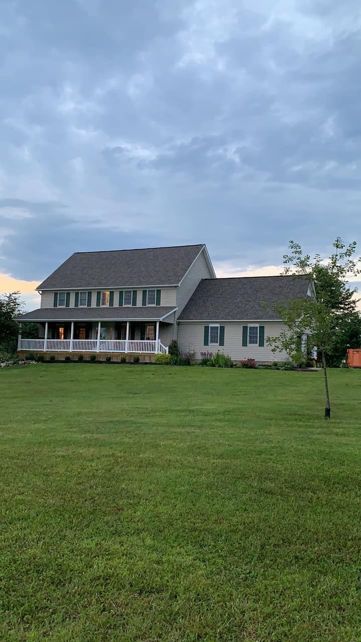 Two-story house with green shutters, a porch, and a gray roof on a grassy lawn under a cloudy sky.