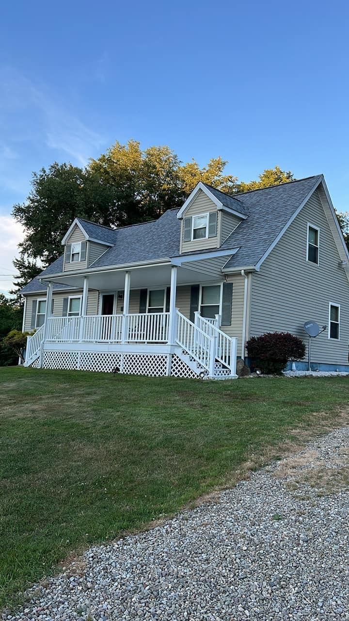 Beige two-story house with white porch and gray roof, situated on a grassy hill under a blue sky.