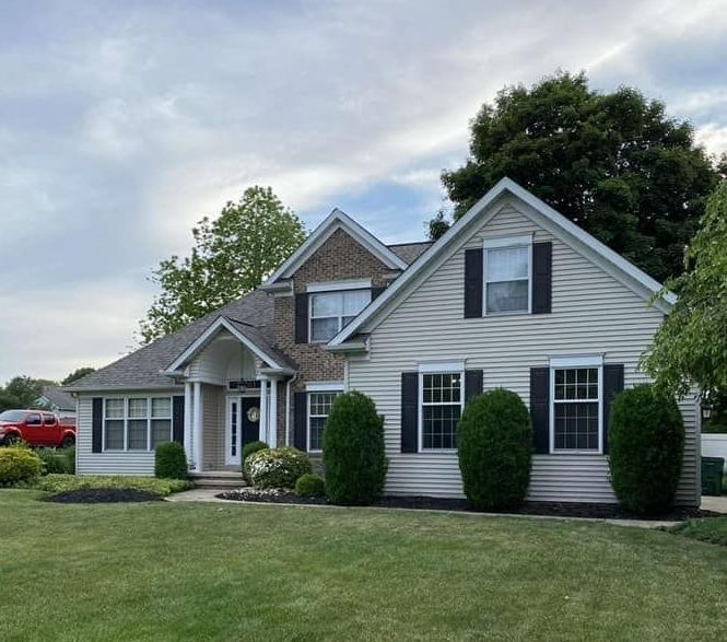 Two-story beige house with black shutters, green lawn, bushes, and a cloudy sky.